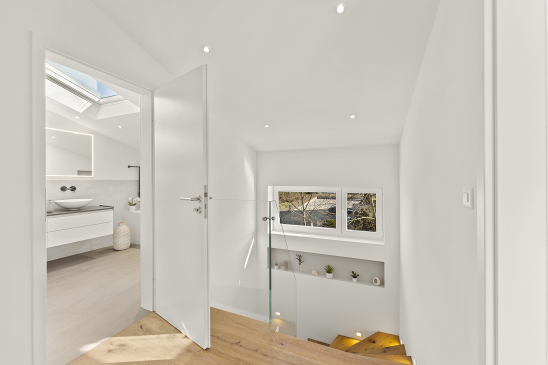 Upper floor hallway in a renovated detached house in Zürich with oak flooring, open door revealing bathroom with vessel sink, glass staircase railing, skylight, and recessed lighting.